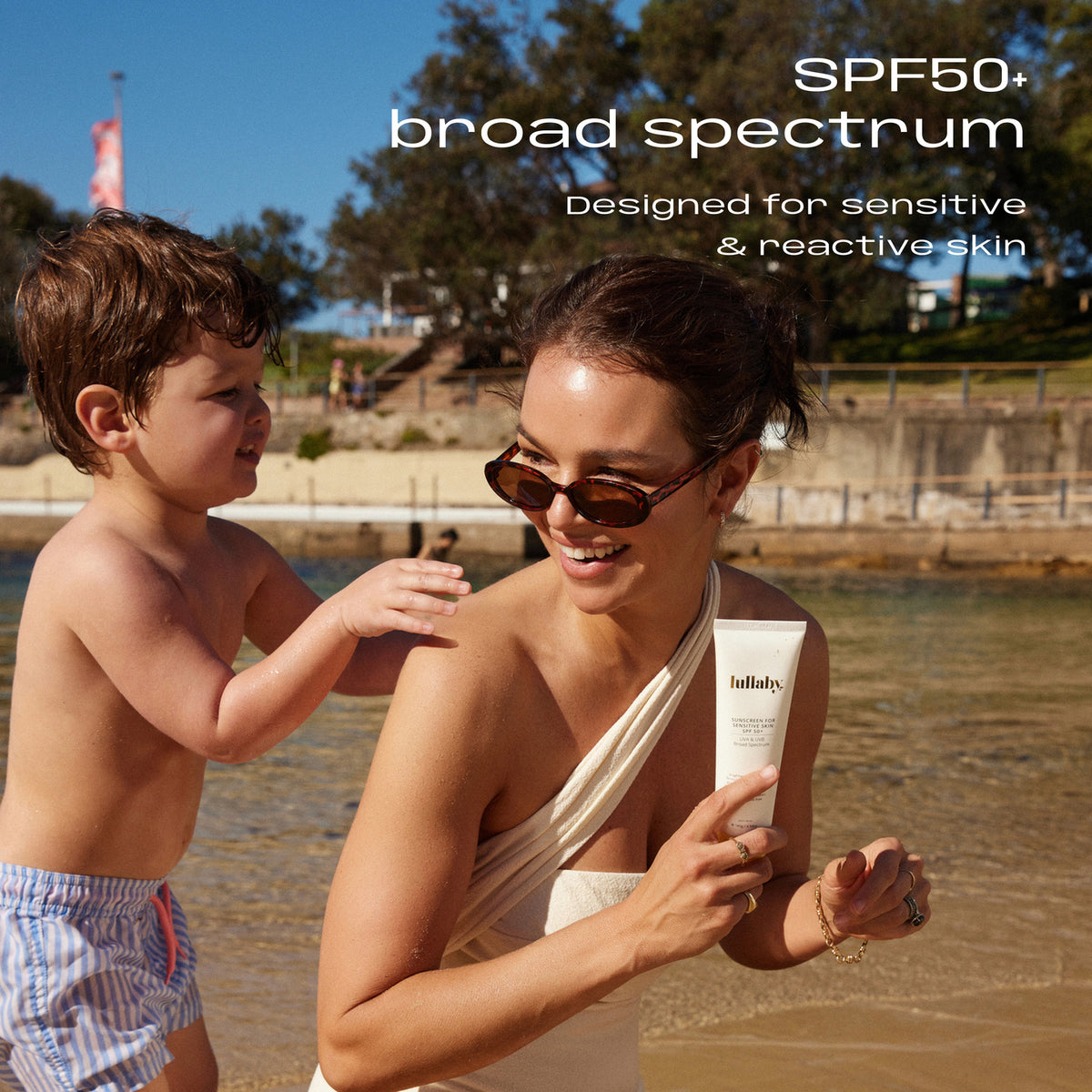 Toddler applying sunscreen to his mum's shoulder at the beach, Lullaby SPF50+ Sensitive Skin Sunscreen 140g in her hand. SPF50+ broad spectrum, designed for sensitive and reactive skin