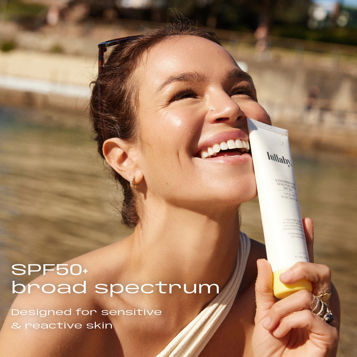 Woman at the beach holding Lullaby SPF50+ Sensitive Skin Sunscreen 140g. SPF50+ broad spectrum, designed for sensitive and reactive skin